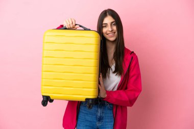 Pembe arka planda izole edilmiş genç beyaz kadın seyahat valiziyle geziyor.