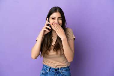 Mor arka planda izole edilmiş cep telefonu kullanan beyaz bir kadın mutlu ve gülümsüyor.