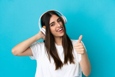 Genç beyaz kadın mavi arka planda izole edilmiş müzik dinliyor ve başparmağını kaldırıyor.