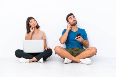 Yerde oturan genç çift beyaz arka planda izole edilmiş bilgisayar ve cep telefonlarını tutarken bir yandan da kafalarını kaşıyarak bir fikir üretiyor.