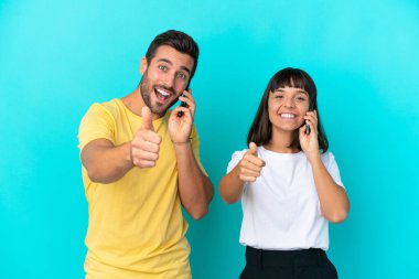 Mavi arka planda izole edilmiş genç bir çift başparmaklarını kaldırırken cep telefonuyla sohbet ediyorlar.
