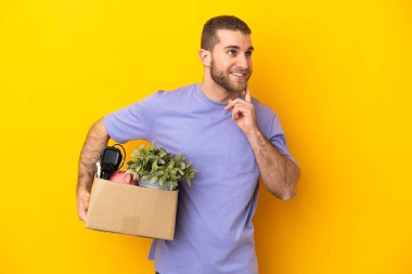 Kafkasyalı genç, sarı arka planda izole edilmiş eşyalarla dolu bir kutuyu kaldırırken yukarı bakarken bir fikir düşünüyor.