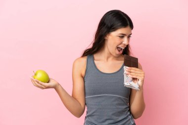 Pembe arka planda izole edilmiş beyaz bir kadın bir elinde çikolata tableti, diğerinde elma alıyor.