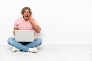 Genç, beyaz arka planda dizüstü bilgisayarıyla yerde oturan beyaz bir adam. Şaşırmış ve şok olmuş bir yüz ifadesi var.