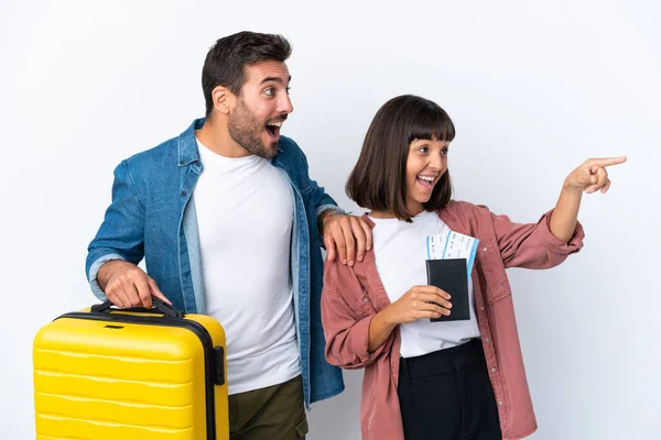 Genç bir çift ellerinde bir bavul ve pasaport ile beyaz arka planda izole edilmiş bir şekilde bir ürün sunmak için kenarı işaret ediyorlar.