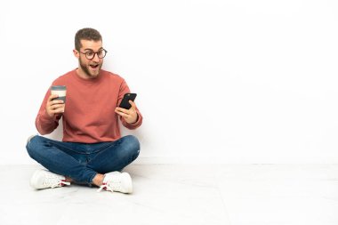 Yerde oturan yakışıklı genç bir adam elinde kahve ve bir cep telefonu tutuyor.