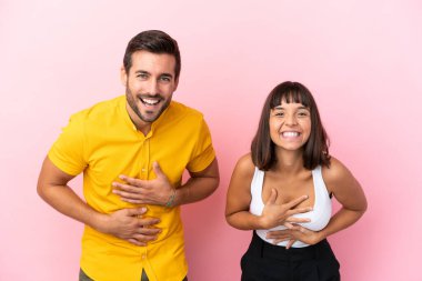 Pembe arka planda yalnız yaşayan genç çift ellerini göğsüne koyup gülümsüyor.