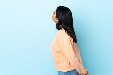 Genç Latin bir kadın mavi arka planda izole edilmiş ve arkasına bakıyor.