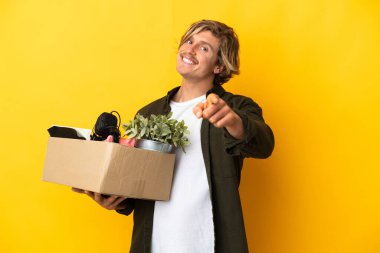 Sarışın adam, sarı arka planda izole edilmiş şeylerle dolu bir kutuyu alıp mutlu bir ifadeyle öne doğru hareket ediyor.