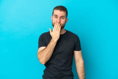 Genç, yakışıklı, beyaz bir adam mavi arka planda izole edilmiş, ağzı elle kapatılmış.