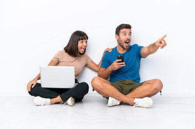 Yerde oturan genç çift, beyaz arka planda izole edilmiş bilgisayar ve cep telefonlarıyla ürünü sunmak için yan tarafa bakıyor.