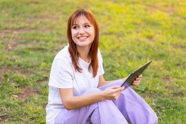 Genç, güzel, kızıl saçlı, açık havada elinde tablet tutan bir kadın.