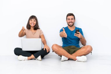 Yerde oturan genç çift beyaz arka planda izole edilmiş cep telefonu ve bilgisayar tutuyor ve iyi bir anlaşma için el sıkışıyorlar.