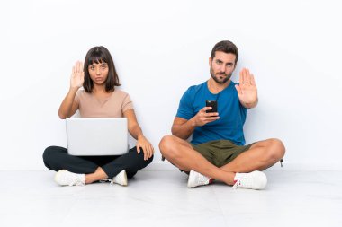 Genç bir çift, beyaz arka planda izole edilmiş bir şekilde ellerinde cep telefonu ve bilgisayar ile yerde oturuyor. Yanlış bir düşünceyi reddeden bir jesti durduruyorlar.