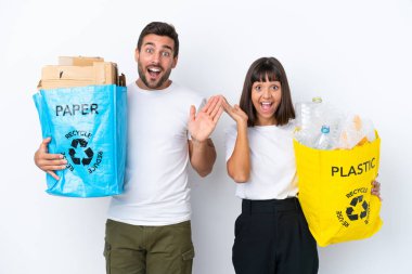 Beyaz arka planda izole edilmiş plastik ve kâğıt dolu bir poşet tutan genç çift şaşırmış ve şok olmuş bir yüz ifadesine sahip.