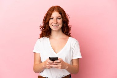 Genç kızılımsı kadın pembe arka planda izole edilmiş kameraya bakıyor ve cep telefonunu kullanırken gülümsüyor.
