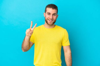 Mavi arka planda izole edilmiş yakışıklı beyaz adam gülümsüyor ve zafer işareti gösteriyor.