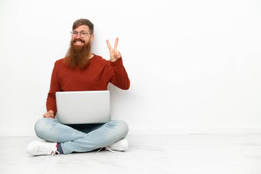 Dizüstü bilgisayarı olan genç, beyaz arka planda izole edilmiş beyaz adam gülümsüyor ve zafer işareti gösteriyor.