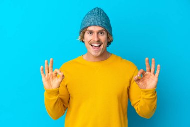 Yakışıklı sarışın adam mavi arka planda izole edilmiş, tamam işareti iki elle gösteriyor.