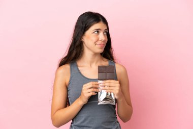 Pembe arka planda izole edilmiş beyaz bir kadın çikolata tableti alıyor ve kuşkuları var.