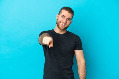 Mavi arka planda izole edilmiş yakışıklı beyaz adam mutlu bir ifadeyle ön tarafı işaret ediyor.