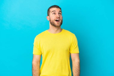 Genç yakışıklı beyaz adam mavi arka planda izole edilmiş yukarı bakıyor ve şaşırmış bir ifadeyle