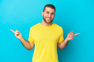 Mavi arka planda izole edilmiş yakışıklı beyaz adam parmak uçlarını işaret ediyor ve mutlu.