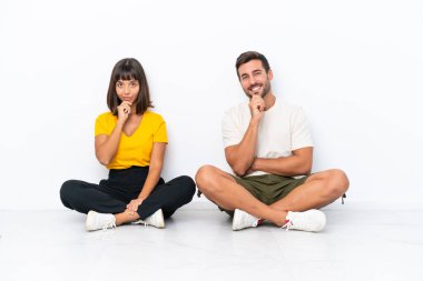 Genç çift beyaz arka planda izole bir şekilde yerde oturuyor gülümsüyor ve kendinden emin bir yüzle öne bakıyor.
