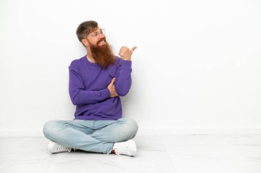 Beyaz arka planda izole bir şekilde yerde oturan beyaz tenli genç beyaz adam bir ürünü sunmak için yan tarafı işaret ediyor.