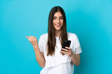Genç beyaz kadın mavi arka planda izole edilmiş cep telefonu kullanıyor ve yan tarafı işaret ediyor.