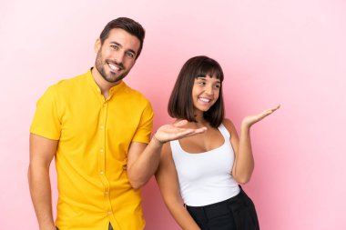 Pembe arka planda izole edilmiş genç çift geriye doğru işaret ediyor ve bir ürün sunuyorlar.