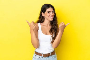 Sarı arka planda izole edilmiş genç beyaz kadın. Baş parmakları havada ve gülümsüyor.