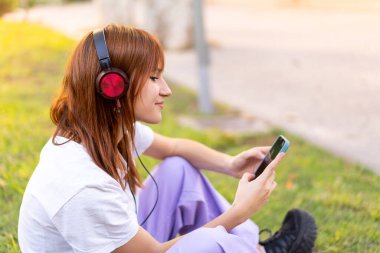 Genç, güzel, kızıl saçlı bir kadın açık havada cep telefonuyla müzik dinliyor.