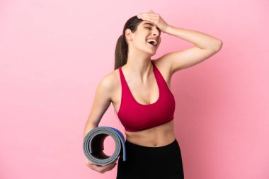 Genç beyaz kadın yoga derslerine giderken elinde minderle gülümsüyor.