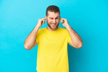 Mavi arka planda izole edilmiş yakışıklı beyaz adam hayal kırıklığına uğramış ve kulaklarını kapatmış.