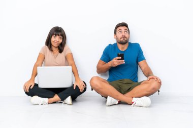 Yerde oturan genç çift, beyaz arka planda izole edilmiş bilgisayar ve cep telefonlarını tutarken, omuzlarını kaldırırken şüpheyle hareket ediyorlar.