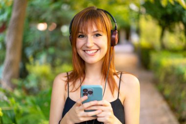 Genç, güzel, kızıl saçlı, açık havada müzik dinleyen, cep telefonu olan ve ön tarafa bakan bir kadın.