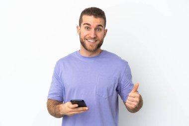 Beyaz arka planda izole edilmiş genç, yakışıklı beyaz bir adam cep telefonu kullanırken baş parmağını kaldırıyor.