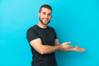 Mavi arka planda izole edilmiş yakışıklı beyaz adam, davet edilmek için ellerini yana uzatıyor.