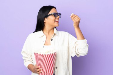 Beyaz arka planda üç boyutlu gözlüklü ve elinde koca bir kova patlamış mısır tutan genç Latin bir kadın.