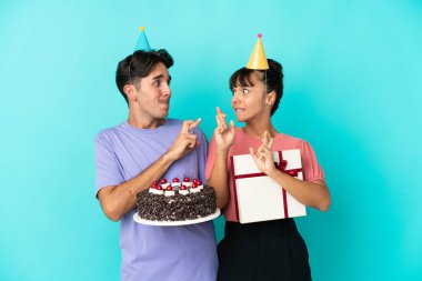 Genç melez bir çift ellerinde doğum günü pastası ve mavi arka planda izole edilmiş bir hediye tutarken şans diliyor.