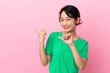 Pembe arka planda izole edilmiş genç Vietnamlı kadın bir ürünü sunmak için kenarı işaret ediyor.