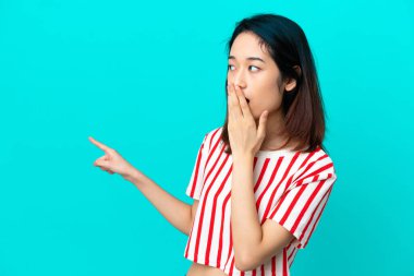 Genç Vietnamlı kadın mavi arka planda izole edilmiş sürpriz bir ifadeyle işaret ederken