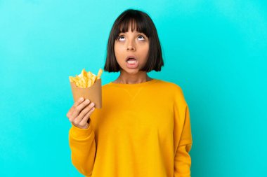 Genç esmer kadın, izole edilmiş mavi arka planda patates kızartması tutuyor ve şaşırmış bir ifadeyle yukarı bakıyor.