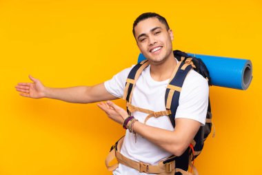 Sarı arka planda izole edilmiş büyük bir sırt çantası olan Asyalı genç adam davet edilmek için ellerini yana uzatıyor.