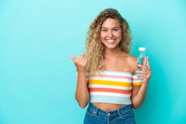 Mavi arka planda izole edilmiş su şişesi olan genç sarışın kadın bir ürünü sunmak için yan tarafı işaret ediyor.