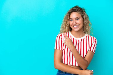 Mavi arka planda yalıtılmış kıvırcık saçlı kız bir ürünü sunmak için kenarı işaret ediyor.