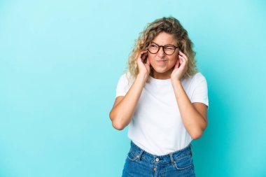 Kıvırcık saçlı kız mavi arka planda izole edilmiş hayal kırıklığına uğramış ve kulaklarını kapatmış.