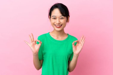 Pembe arka planda izole edilmiş genç Vietnamlı kadın iki eliyle tamam işareti gösteriyor.