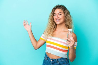 Mavi arka planda izole edilmiş su şişesi olan genç sarışın kadın davet etmek için ellerini yana uzatıyor.